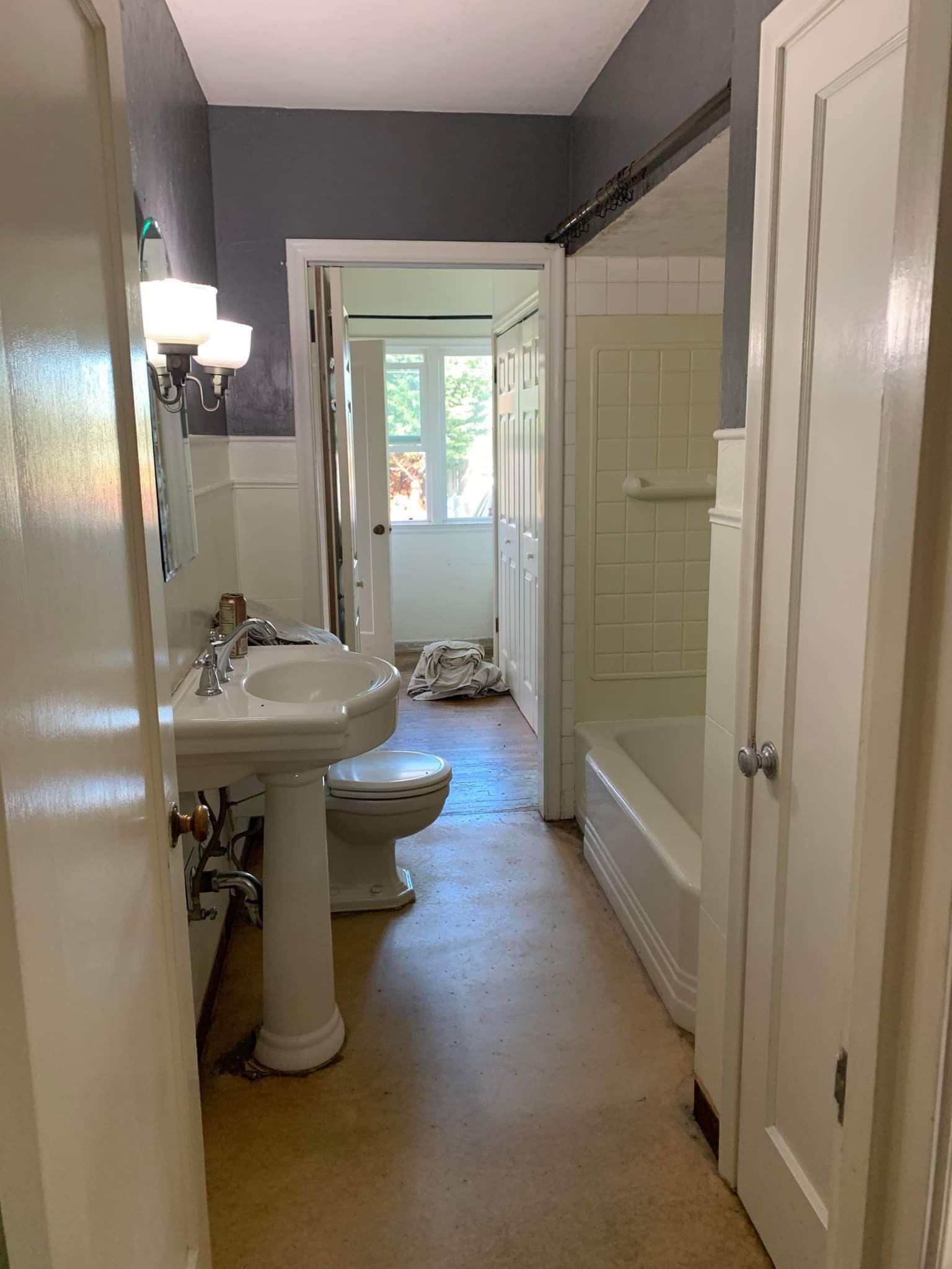 Bathroom with white fixtures, grey walls, and a view into a small room with natural light.