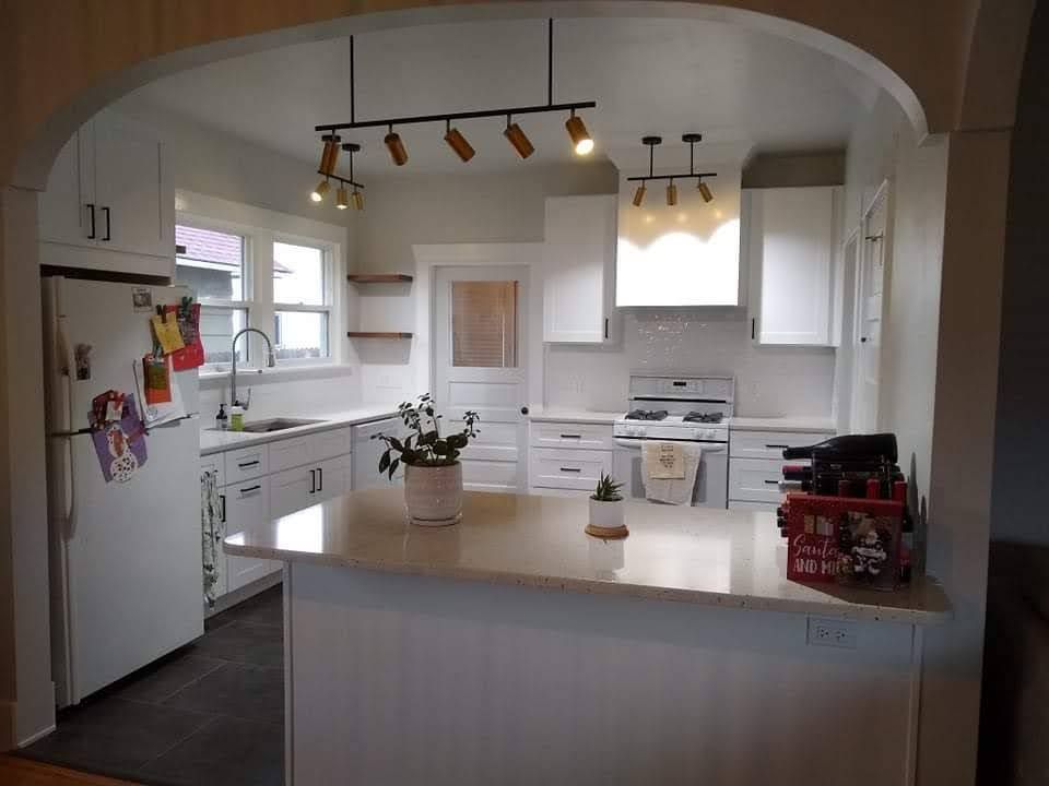 White kitchen with an island, cabinets, and appliances. It has a light-colored countertop, and modern light fixtures.