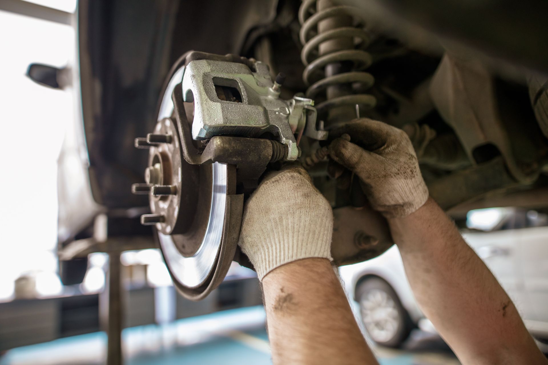 Mechanic working on a car brake assembly under the vehicle, wearing gloves.