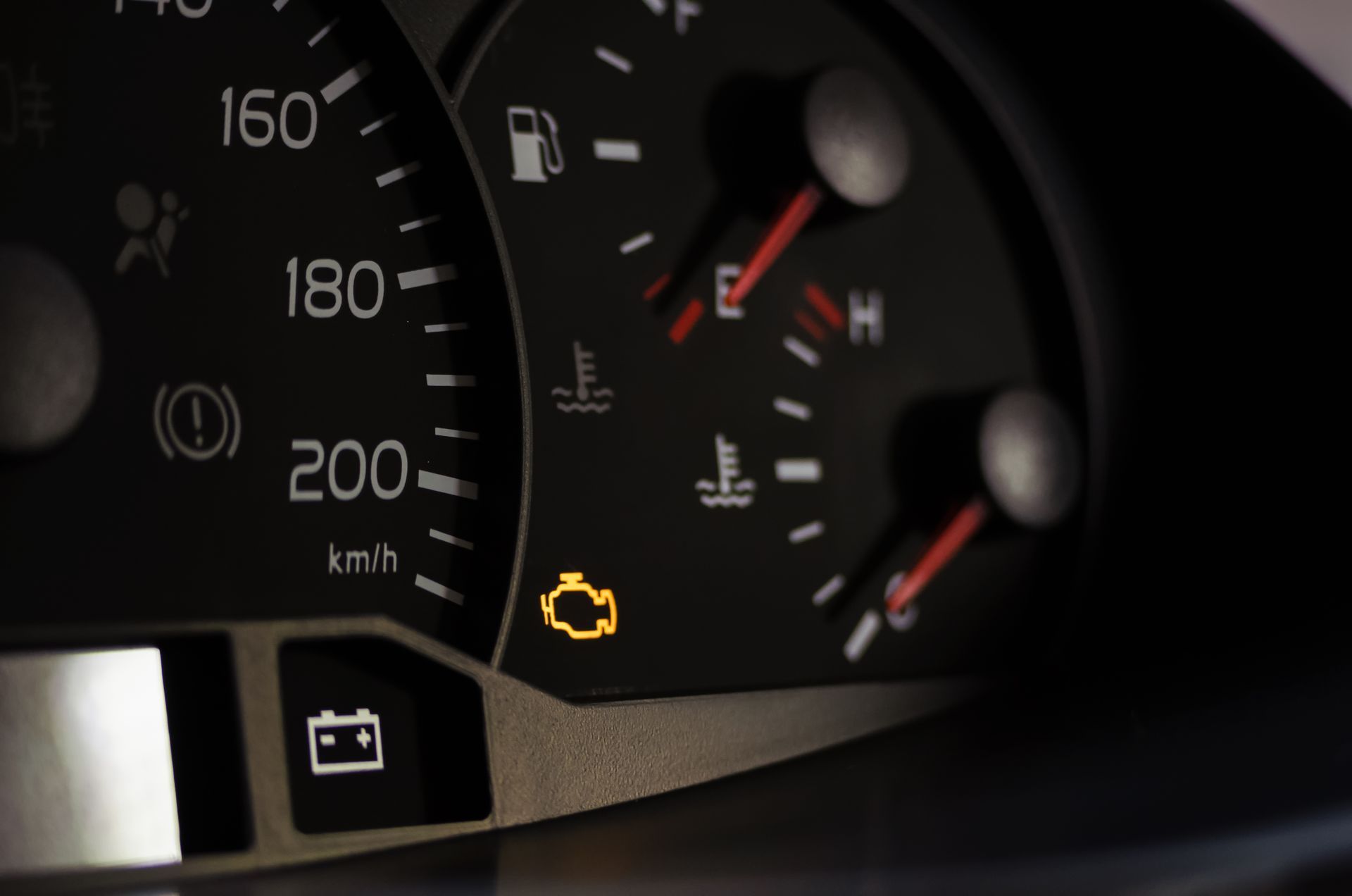 Car dashboard gauges with a glowing yellow check engine warning light and battery icon.