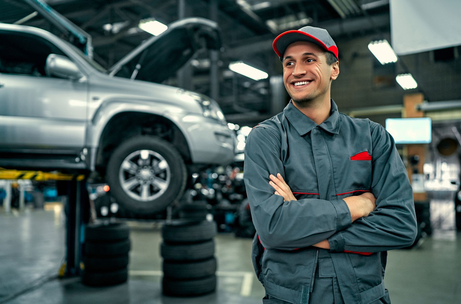 Mechanic smiling in a garage beside a lifted silver SUV on a car jack