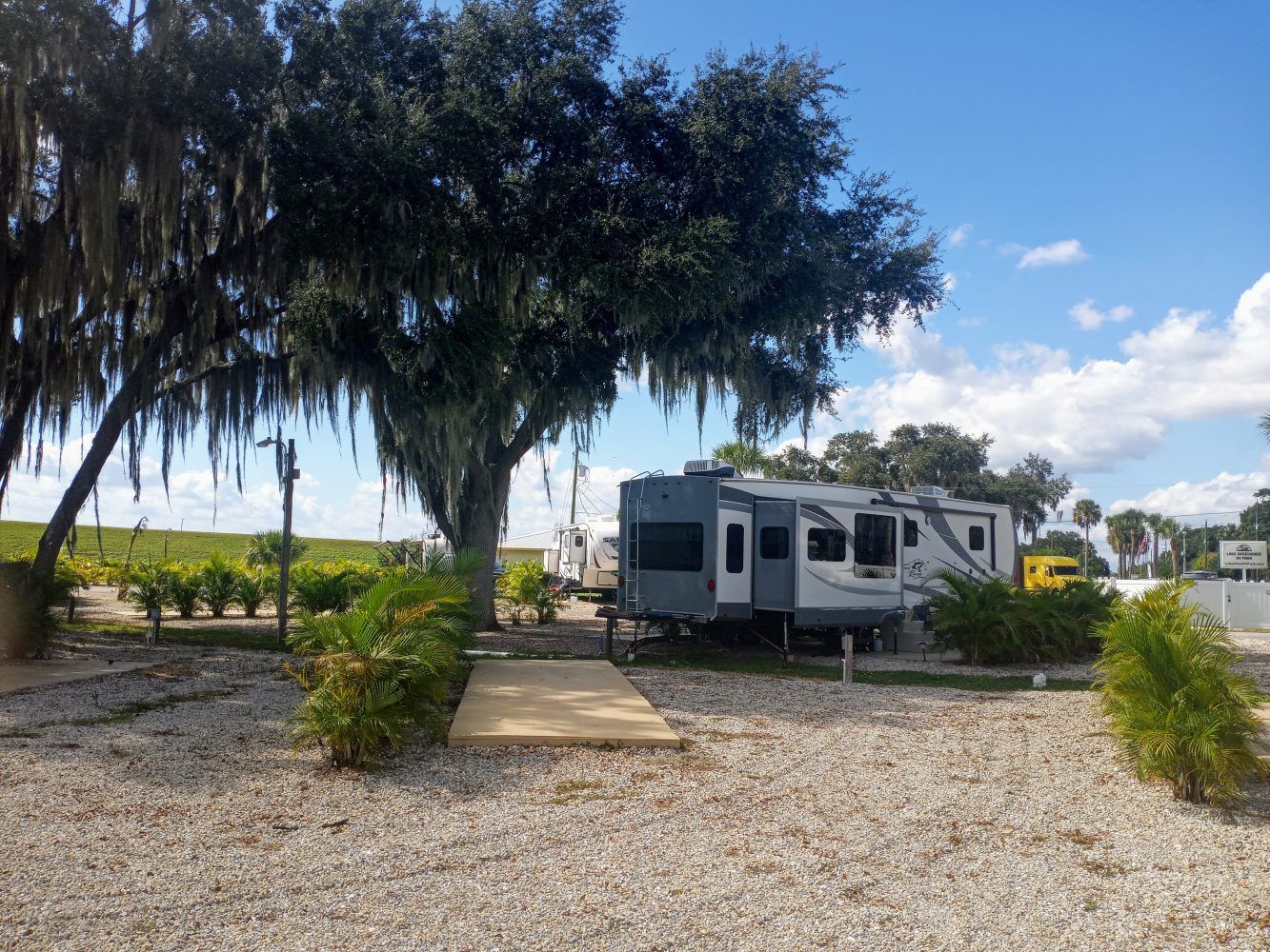 RV parked on gravel in a campground.  Green bushes and tree in view, blue sky.