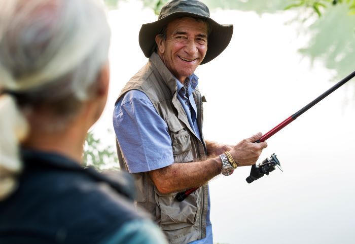 Smiling man fishing with woman, wearing a hat and vest outdoors.