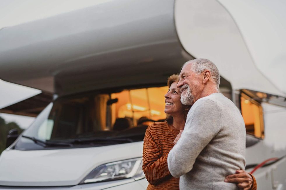 Elderly couple hugging, smiling in front of a white RV; outdoor setting, sunset glow.