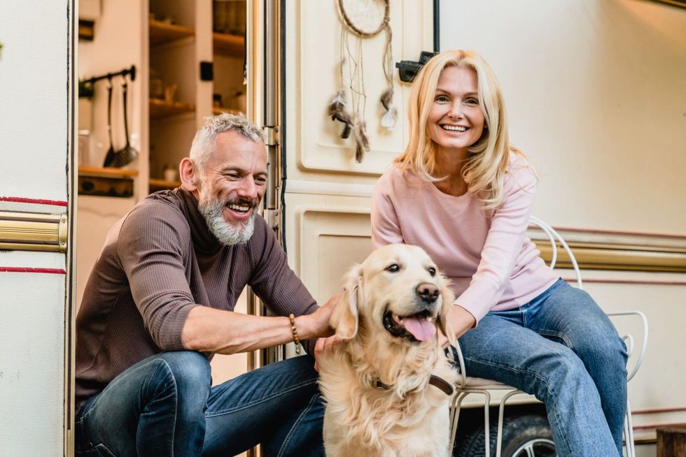 Smiling couple with a Golden Retriever dog by a camper. They sit outdoors, the woman petting the dog.