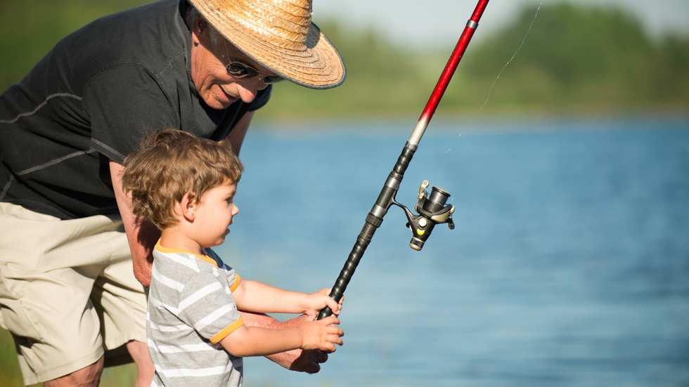Grandfather teaching a young child to fish by a lake, holding a fishing rod together.