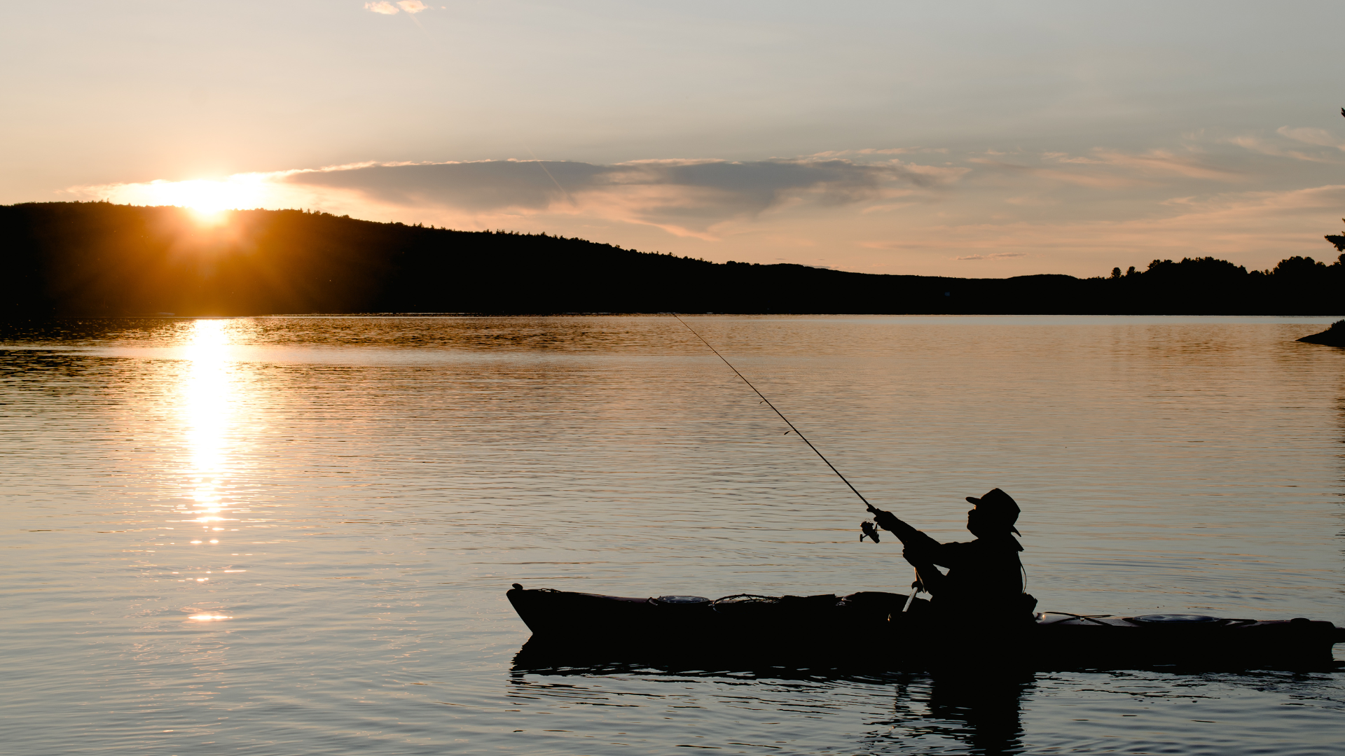 Silhouette of person fishing from kayak at sunset over lake.