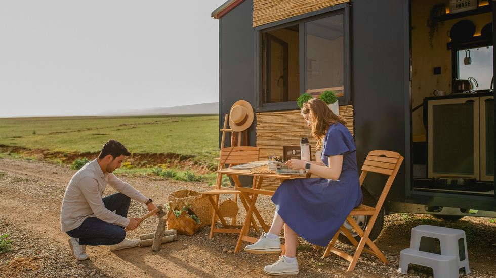 Man and woman near a tiny house; man hammering, woman seated at table eating, outdoor setting.