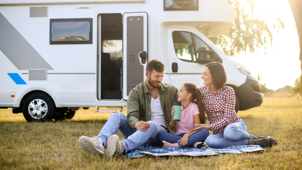 Family picnicking on a blanket in front of a campervan at sunset; smiling, happy.