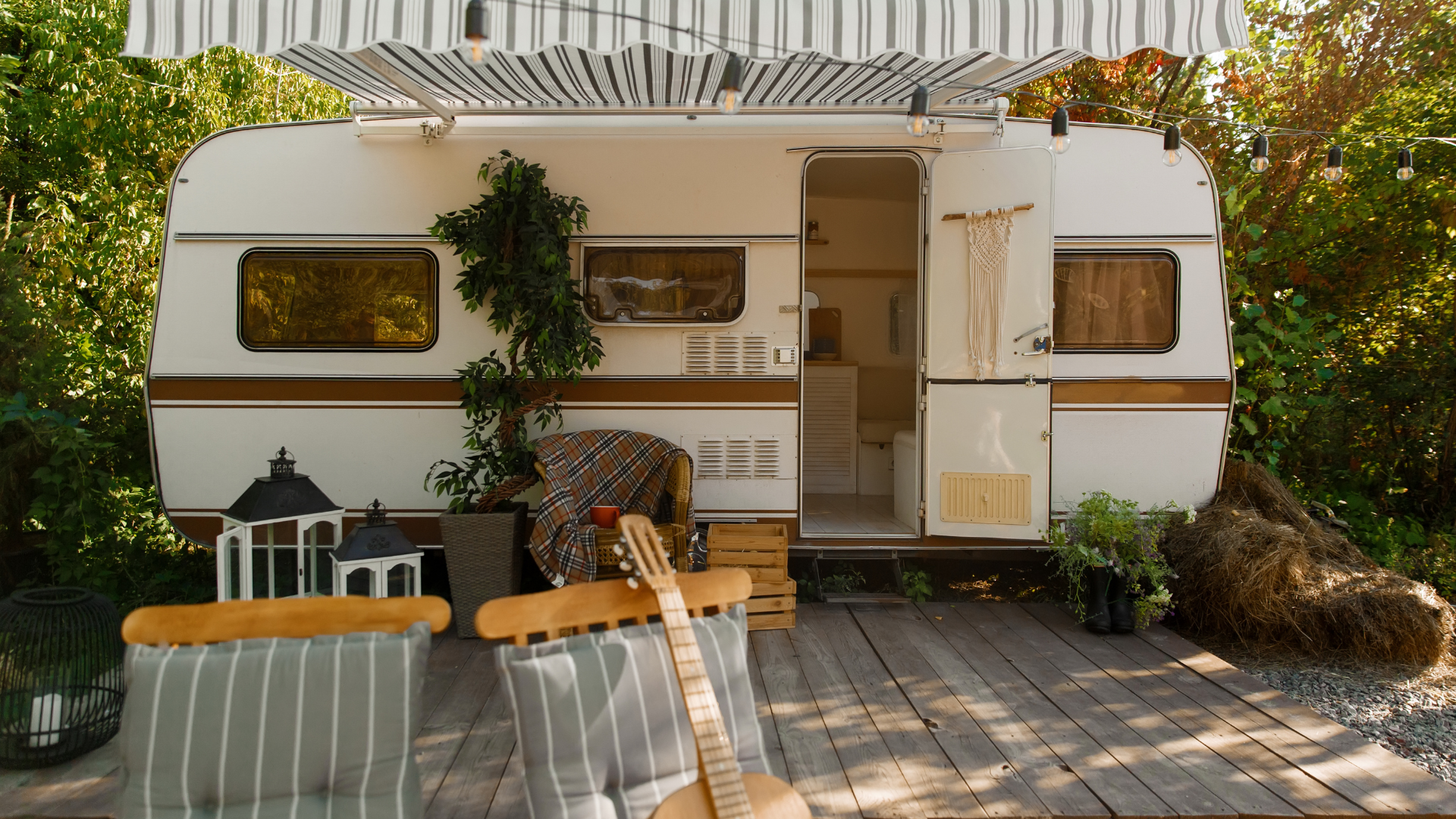 White camper with awning and open door on wooden deck, surrounded by plants and chairs; guitar leaning nearby.