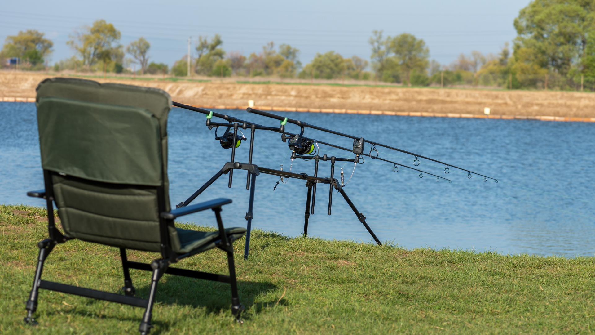 Green folding chair by lake; fishing rods on stand, sunny day.