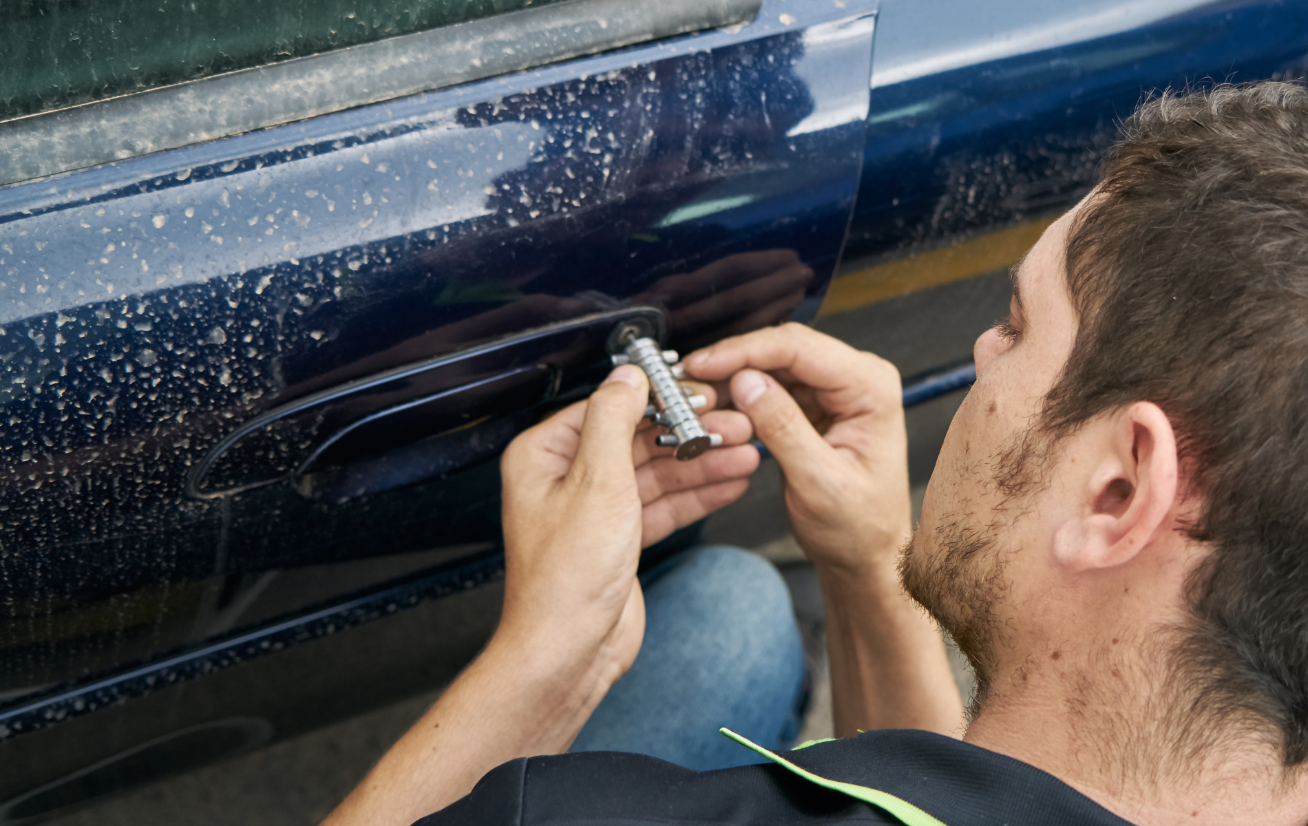 A person uses a specialized automotive tool to repair the lock on the driver-side door of a dark blue vehicle.