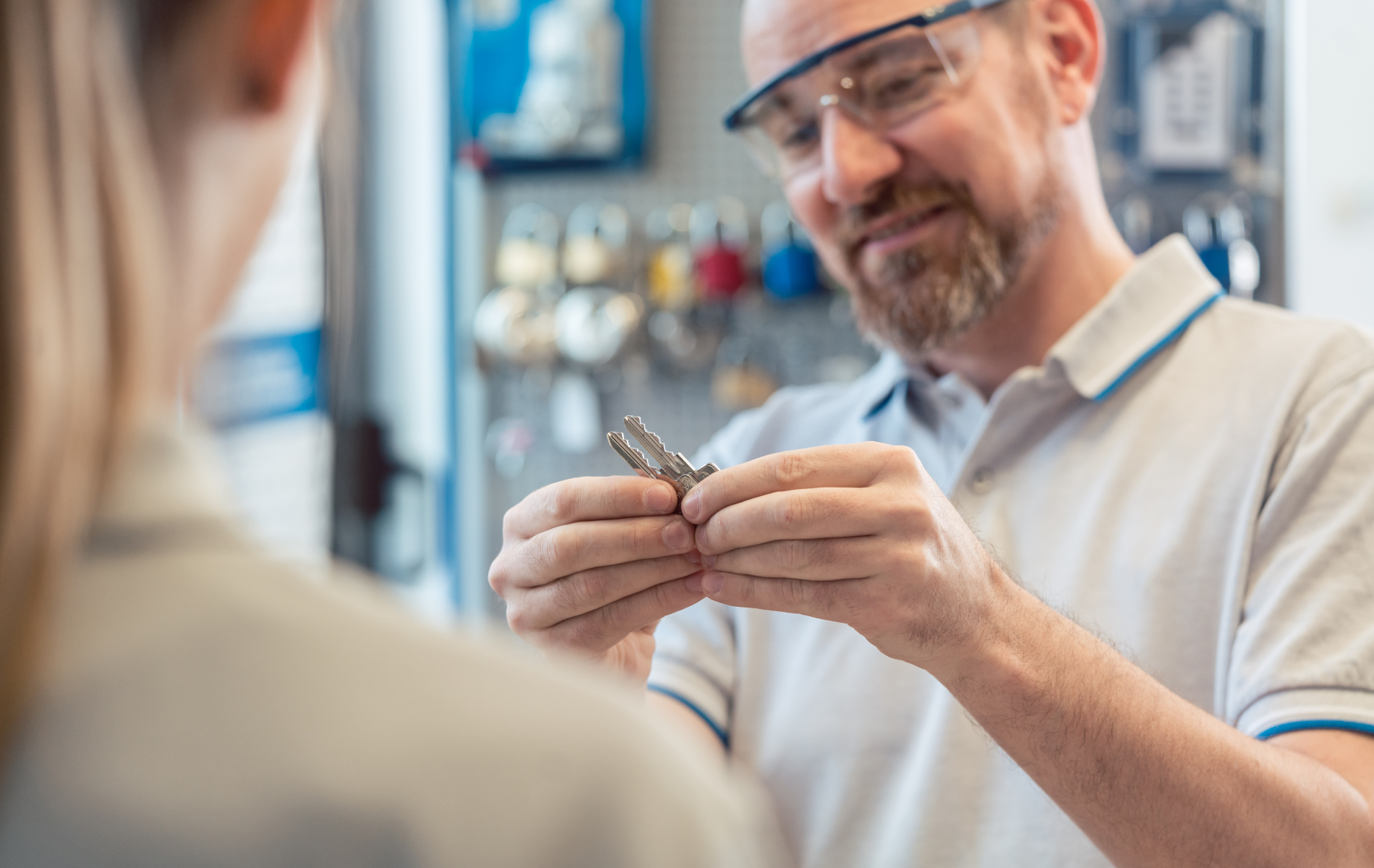 Man in safety glasses examines keys while speaking to a person, shop background.