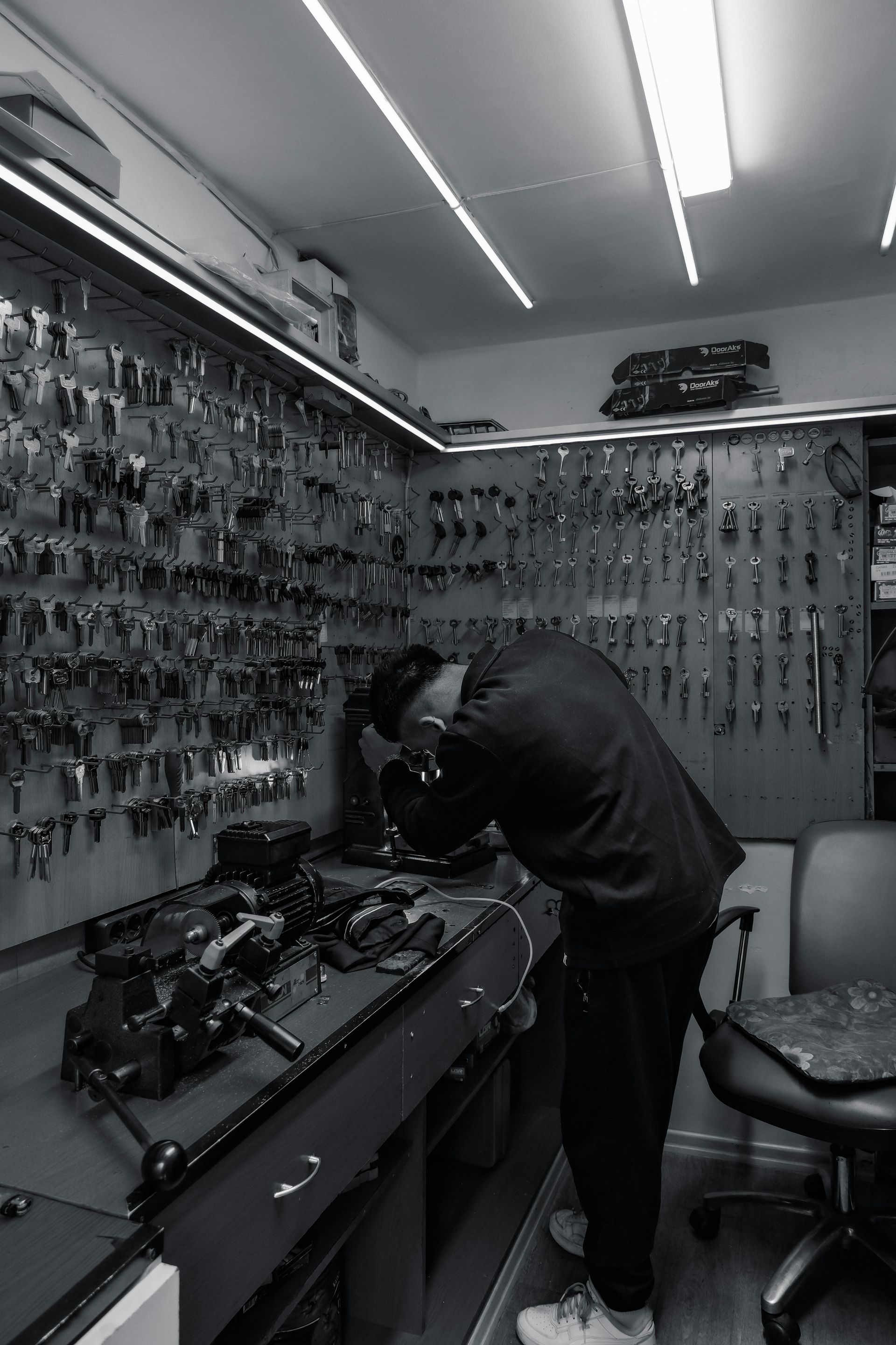 Locksmith working on keys in shop, with rows of keys displayed on wall and workbench.