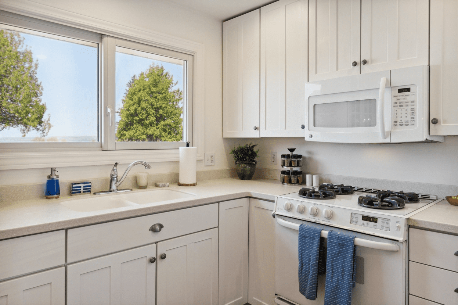 A kitchen with white cabinets , a stove , a microwave , a sink and a window.