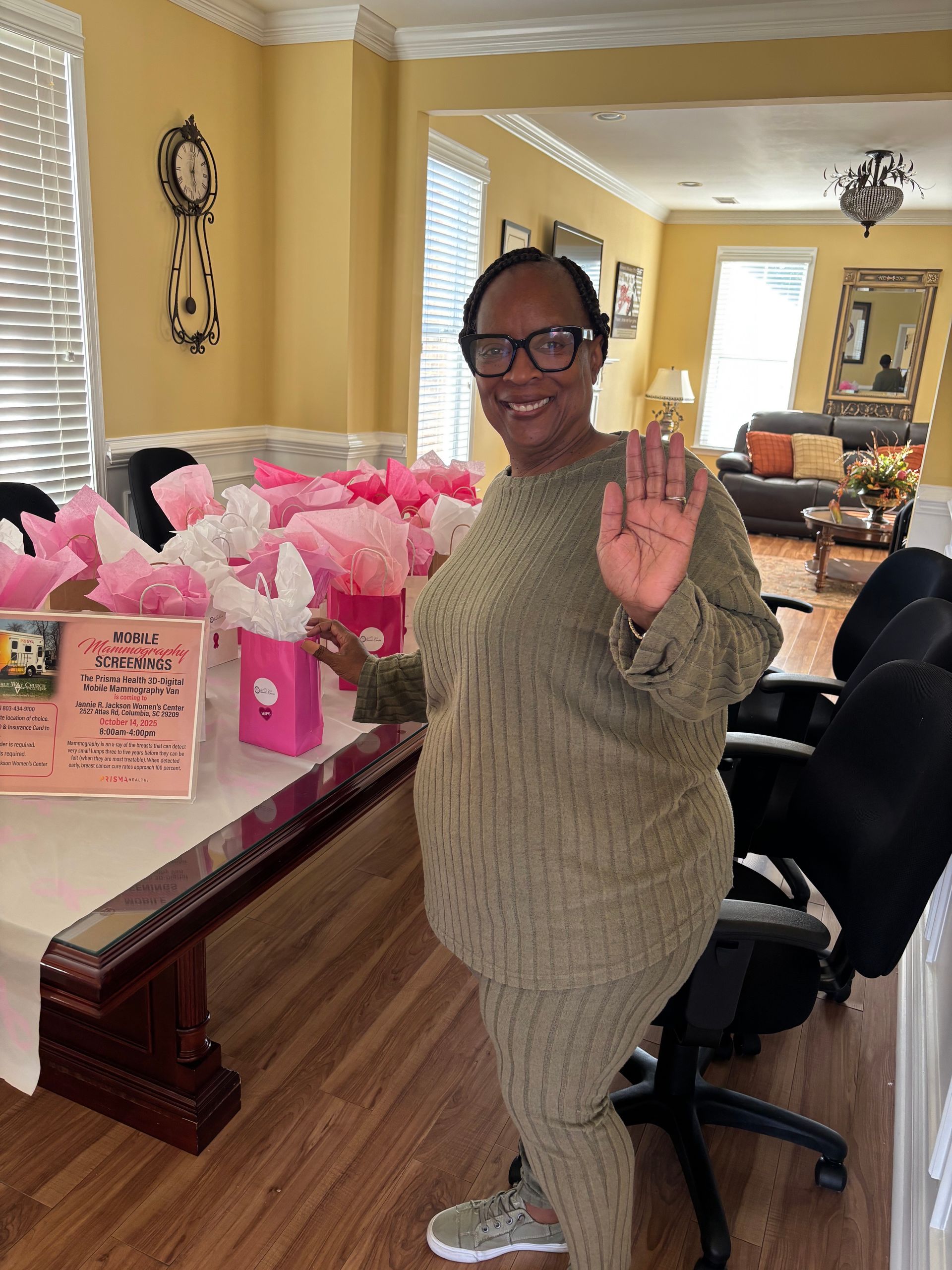 Woman in olive green outfit waving, standing near a table with pink decorations and a sign.