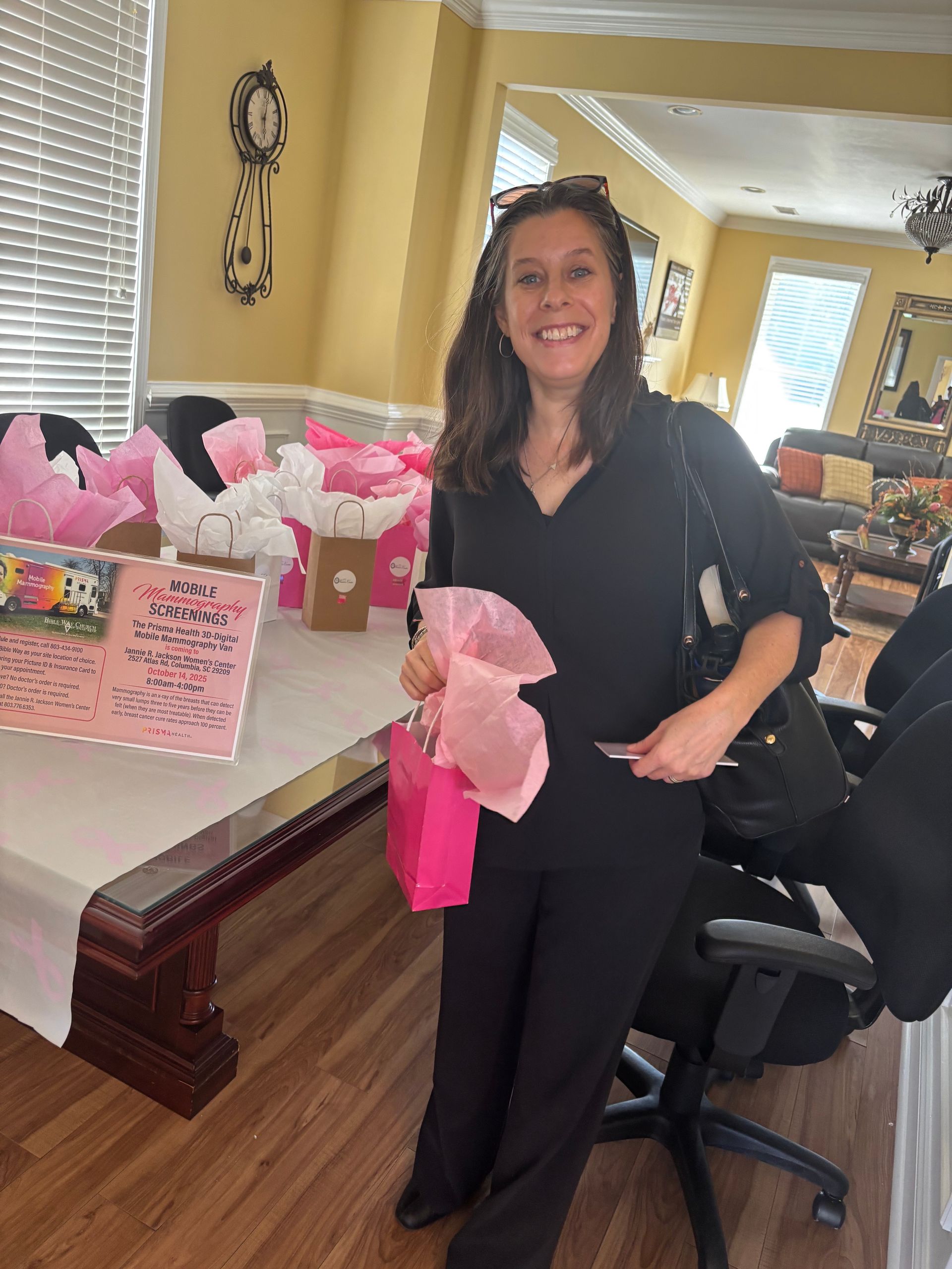 Woman smiles, holding pink gift bag. Standing near table with more gifts, in decorated room.