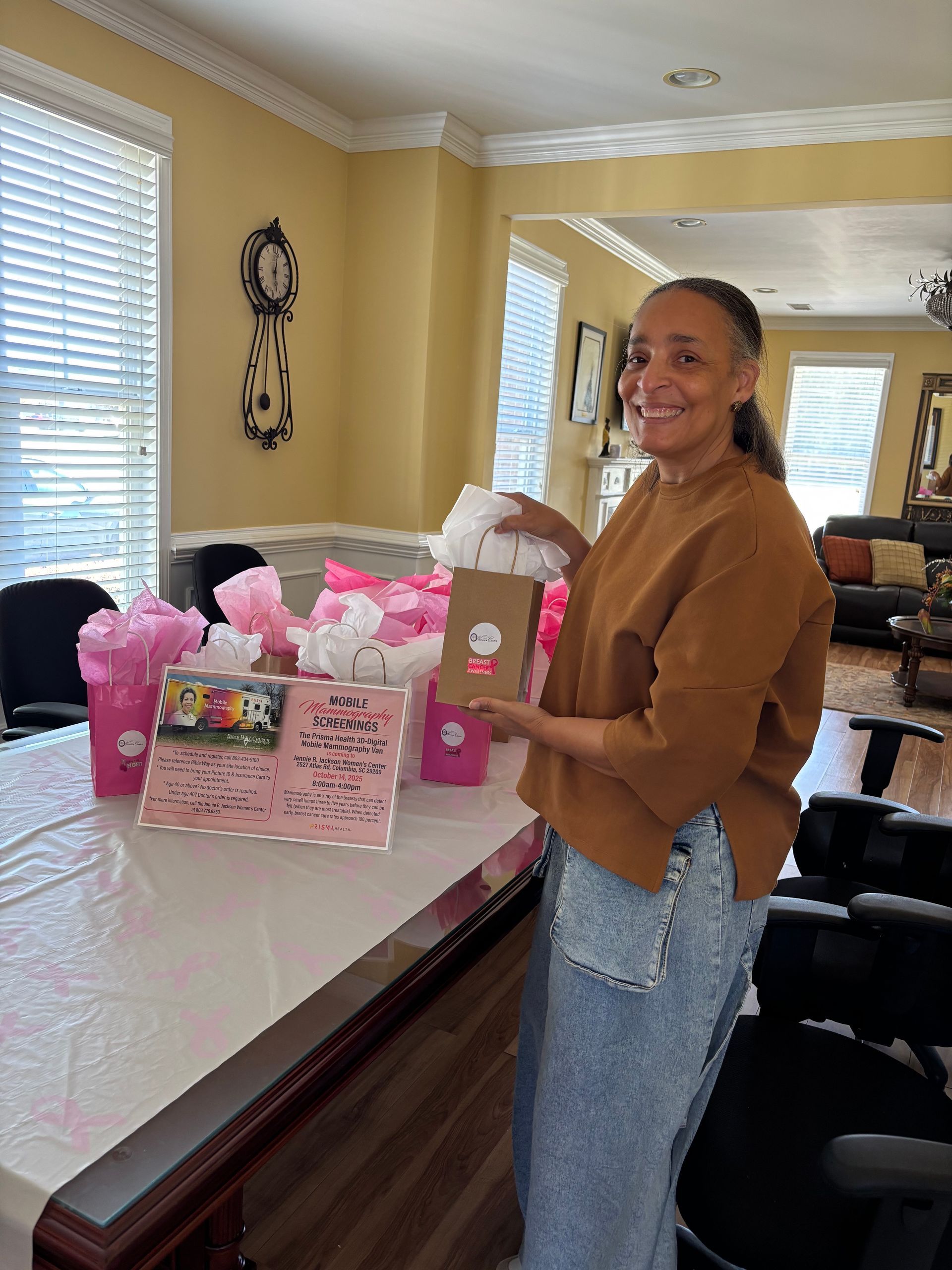 Woman holds gift, smiling, by a table with pink decorations, indoors with sunny windows.