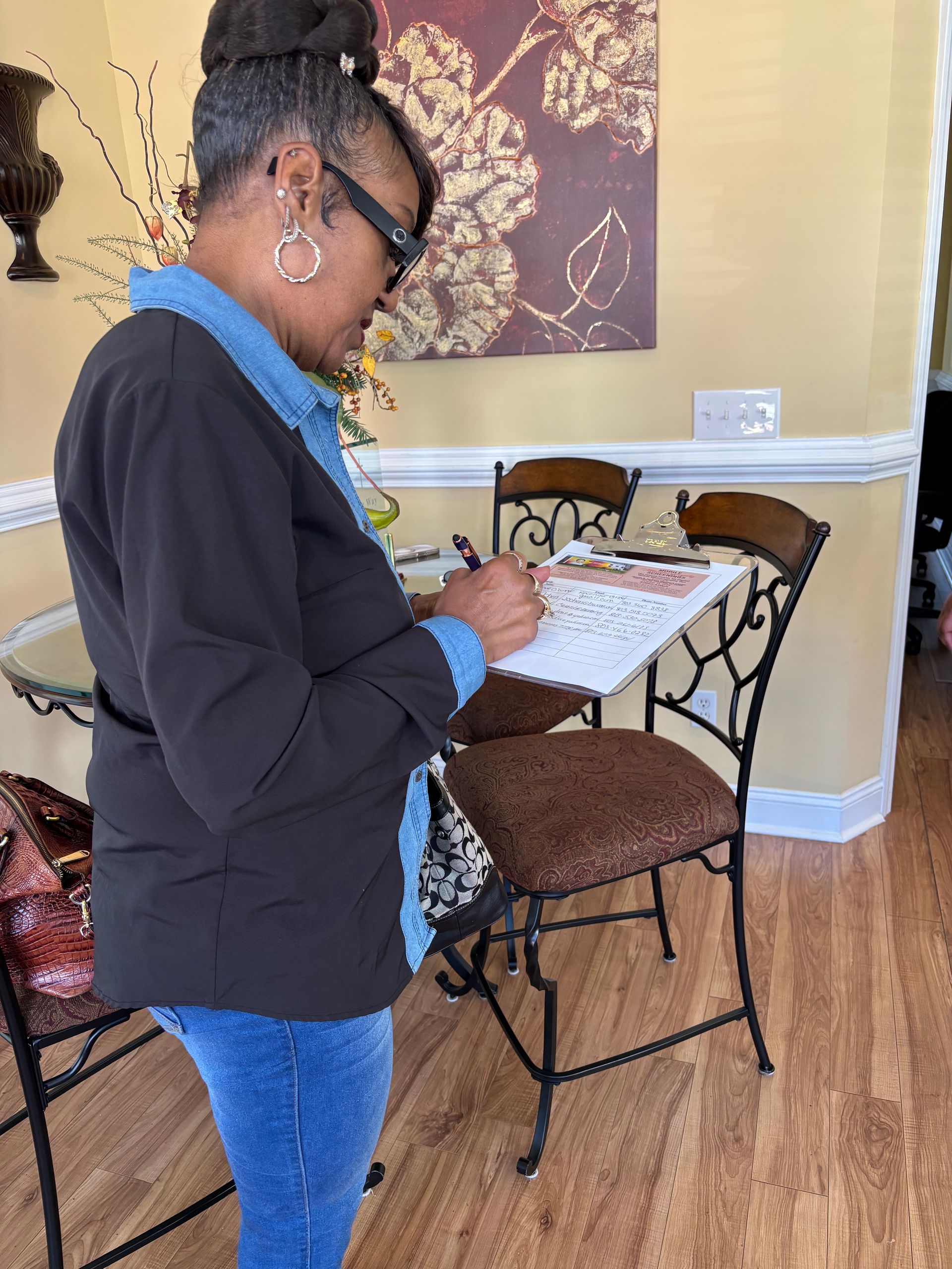 Woman writing on a clipboard in a dining area; wearing glasses, jeans, and a jacket.