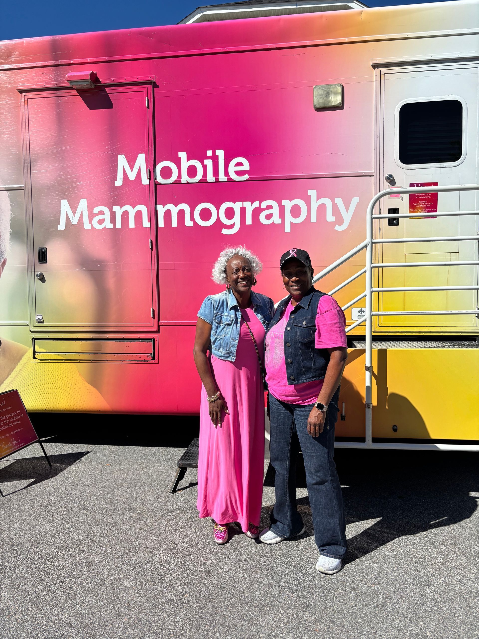 Two women stand in front of a mobile mammography unit. The unit is pink and orange.