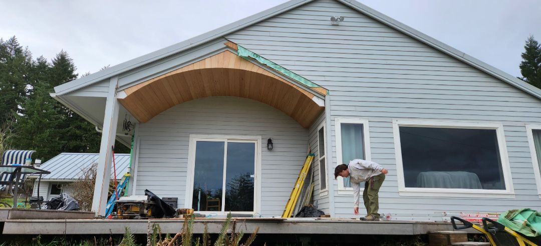 A person working on a house porch under construction. The house has gray siding and an arched wooden soffit.