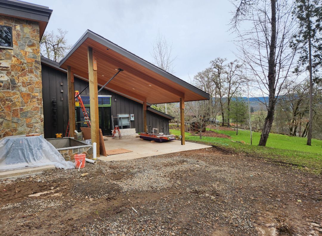 A wooden-framed carport with a concrete slab extends from a dark-colored building near a grassy hillside.