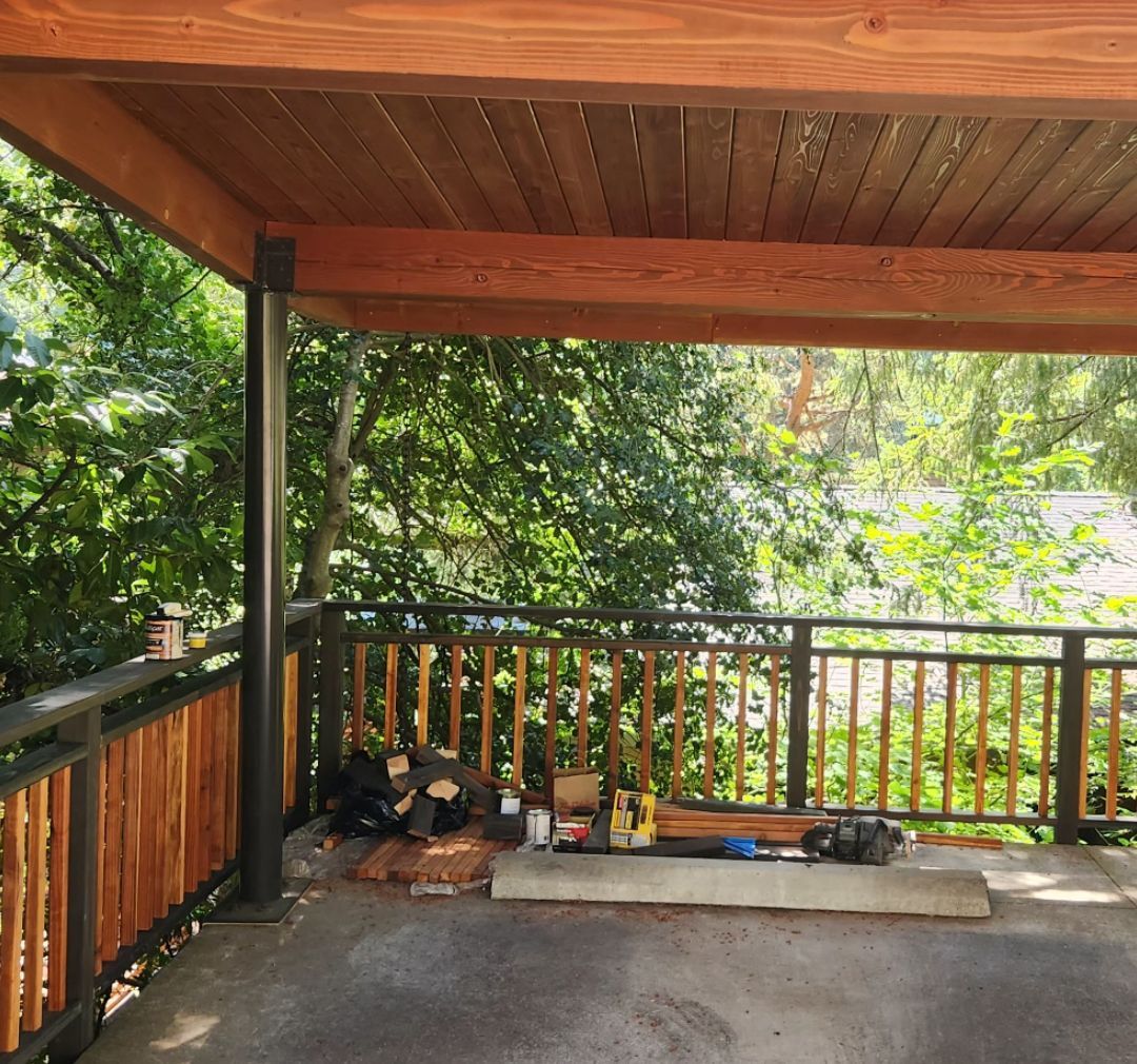 Covered outdoor deck with wood railings, overlooking lush greenery and a body of water.