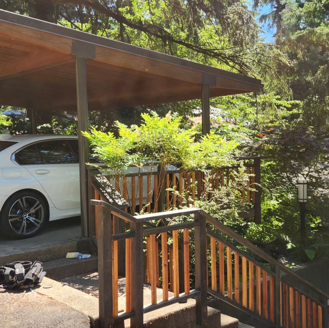 Car under a carport next to wooden stairs and a lush green landscape.