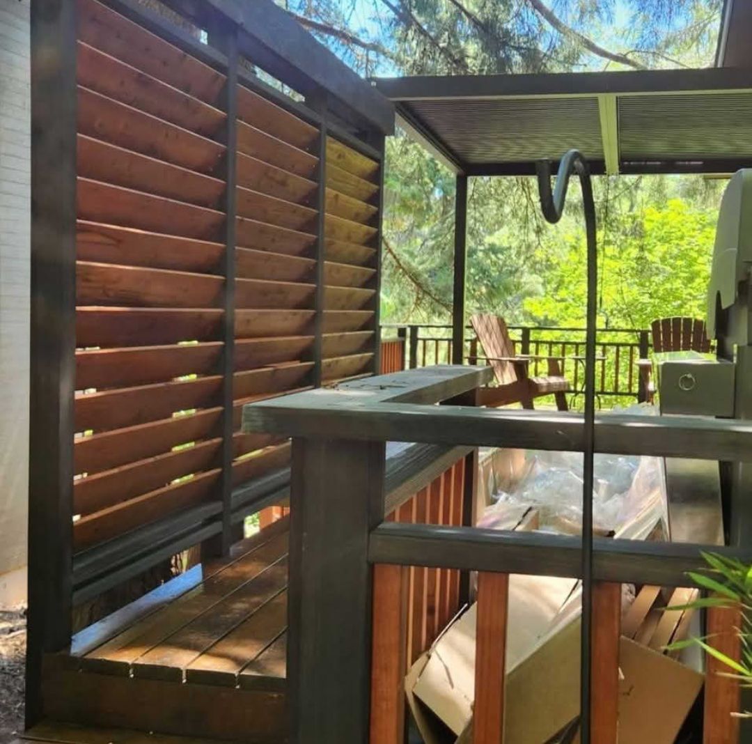 Wooden slatted privacy screen on a deck with a grill and outdoor furniture, trees in the background.