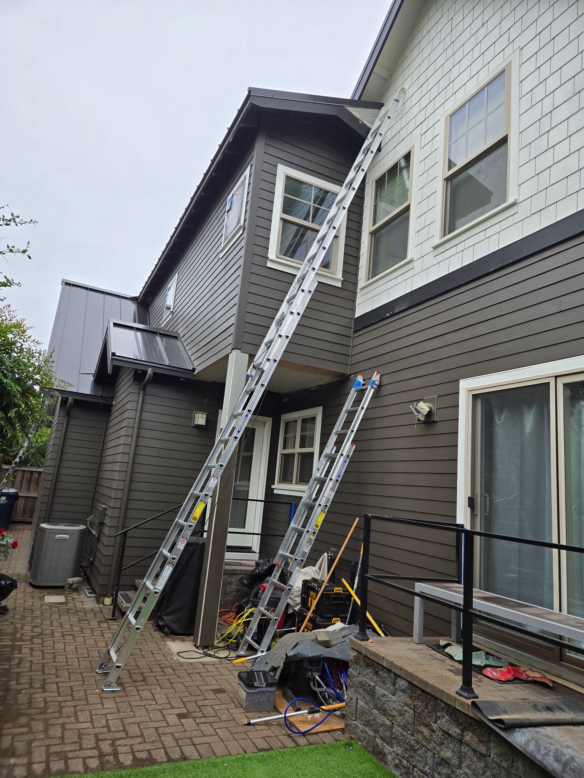 Ladders propped against a two-story house with dark brown siding; construction tools are on the ground.