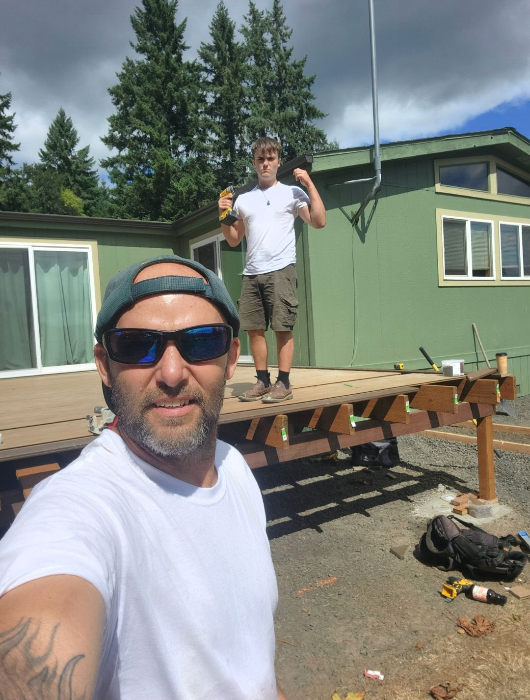Two men on a deck; one takes a selfie, the other stands. Green building and trees in the background.