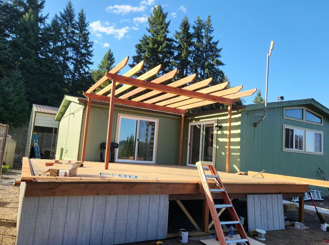 Wooden deck under construction with a pergola attached to a green house, sunny day.