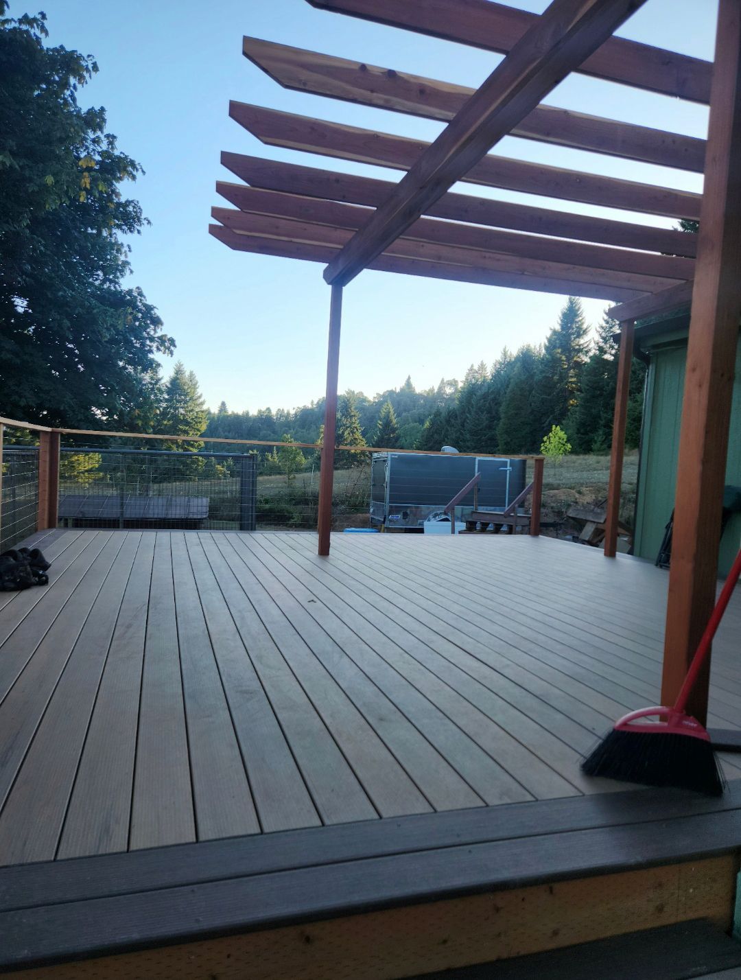 Wooden deck with pergola, overlooking a forested landscape under a clear sky. A broom leans against the house.