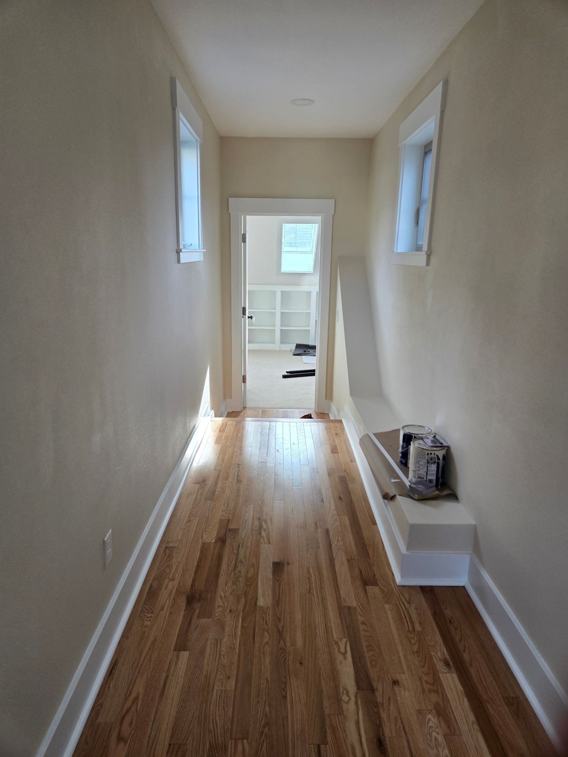 Narrow hallway with hardwood floor, off-white walls, two windows, and door to a room.