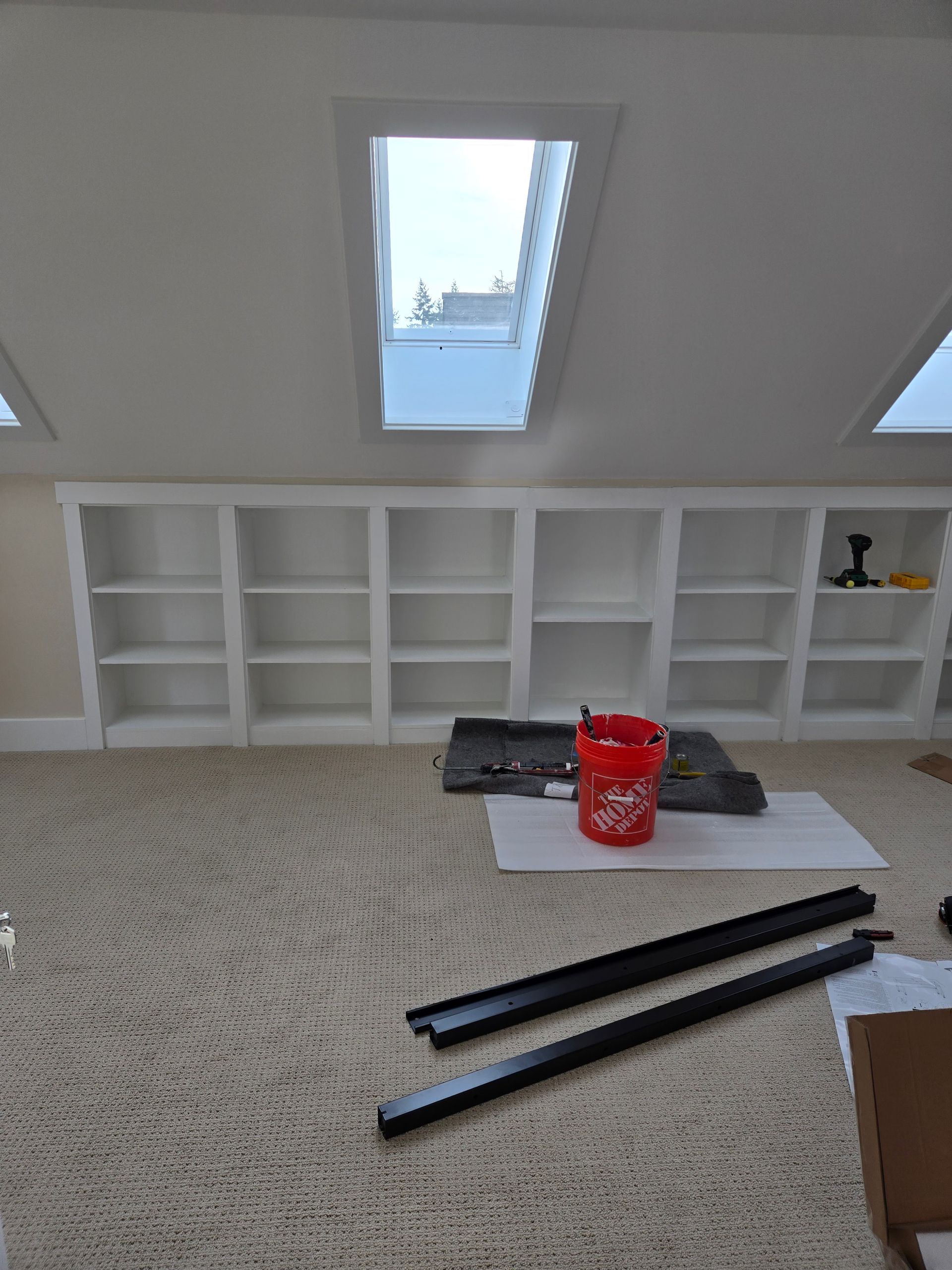 Built-in white bookshelves on a beige carpet, with a skylight above. A red bucket sits in the center.