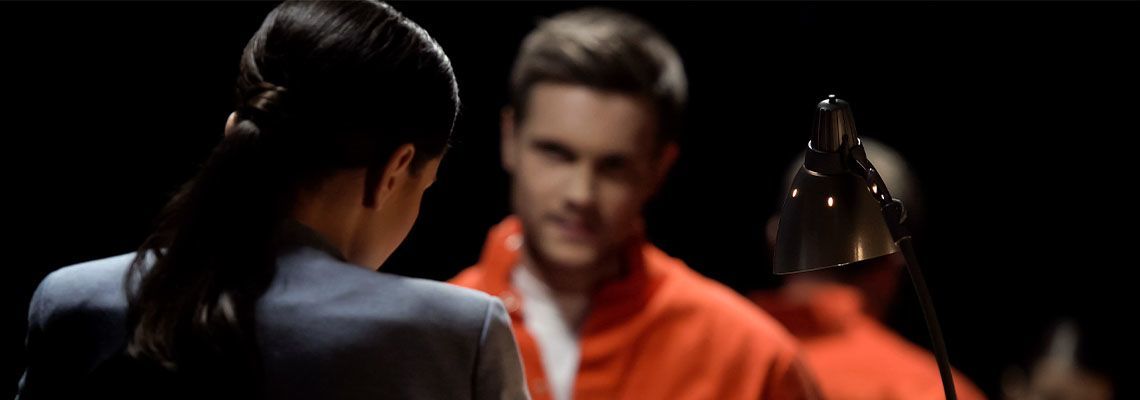 A woman in a blue suit faces a man in an orange jumpsuit. The man smiles. Dark background.