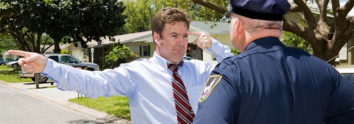 A man gesturing and pointing, speaking to a police officer outdoors in front of houses and trees.