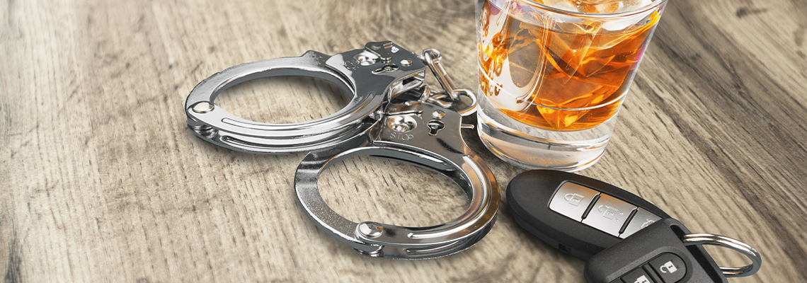 Handcuffs, glass of whiskey, and car keys on a wooden surface, suggesting drunk driving.