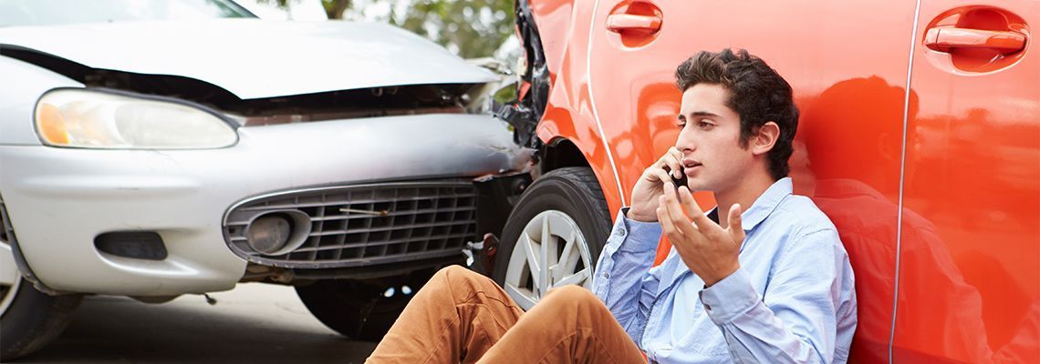 A man sits near a red car, on the phone. Damaged cars are in the background, likely a car crash scene.