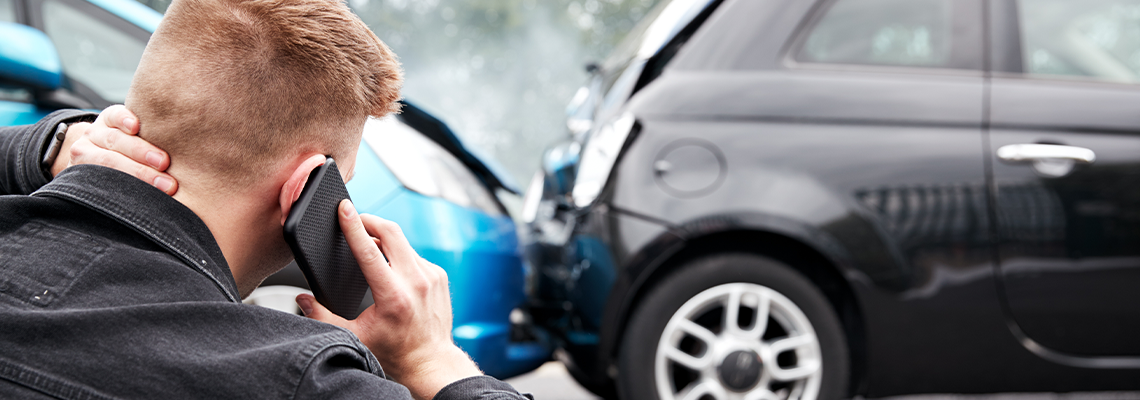 Man on phone at a car accident, holding his neck, with a blue and black car involved.