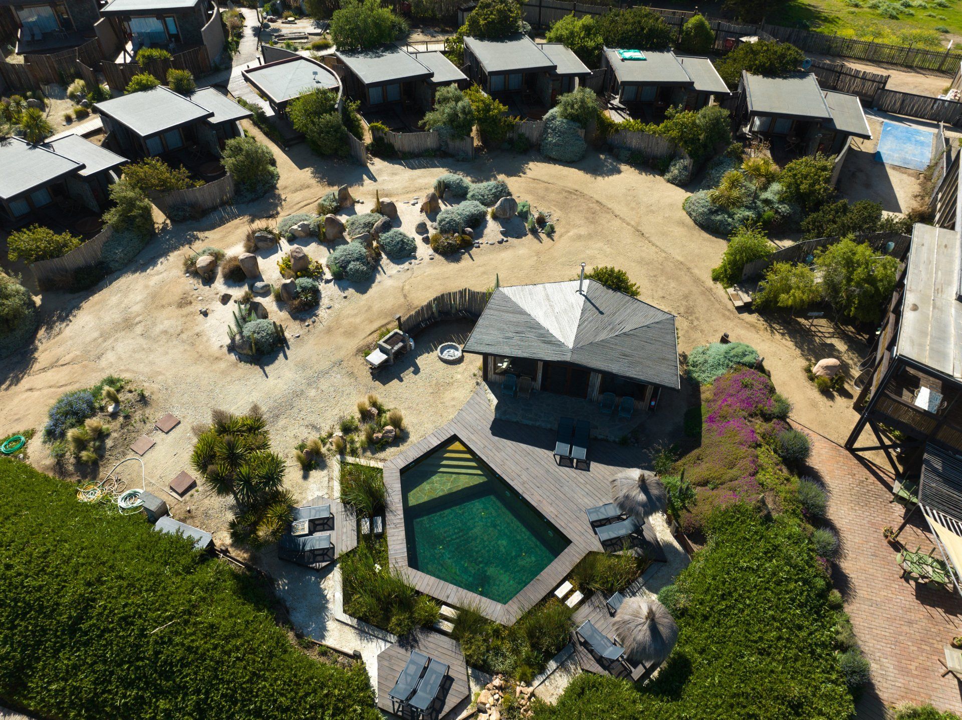 Aerial view of a resort with small cabins, a pool, and a central pavilion with a dark roof and surrounding greenery.