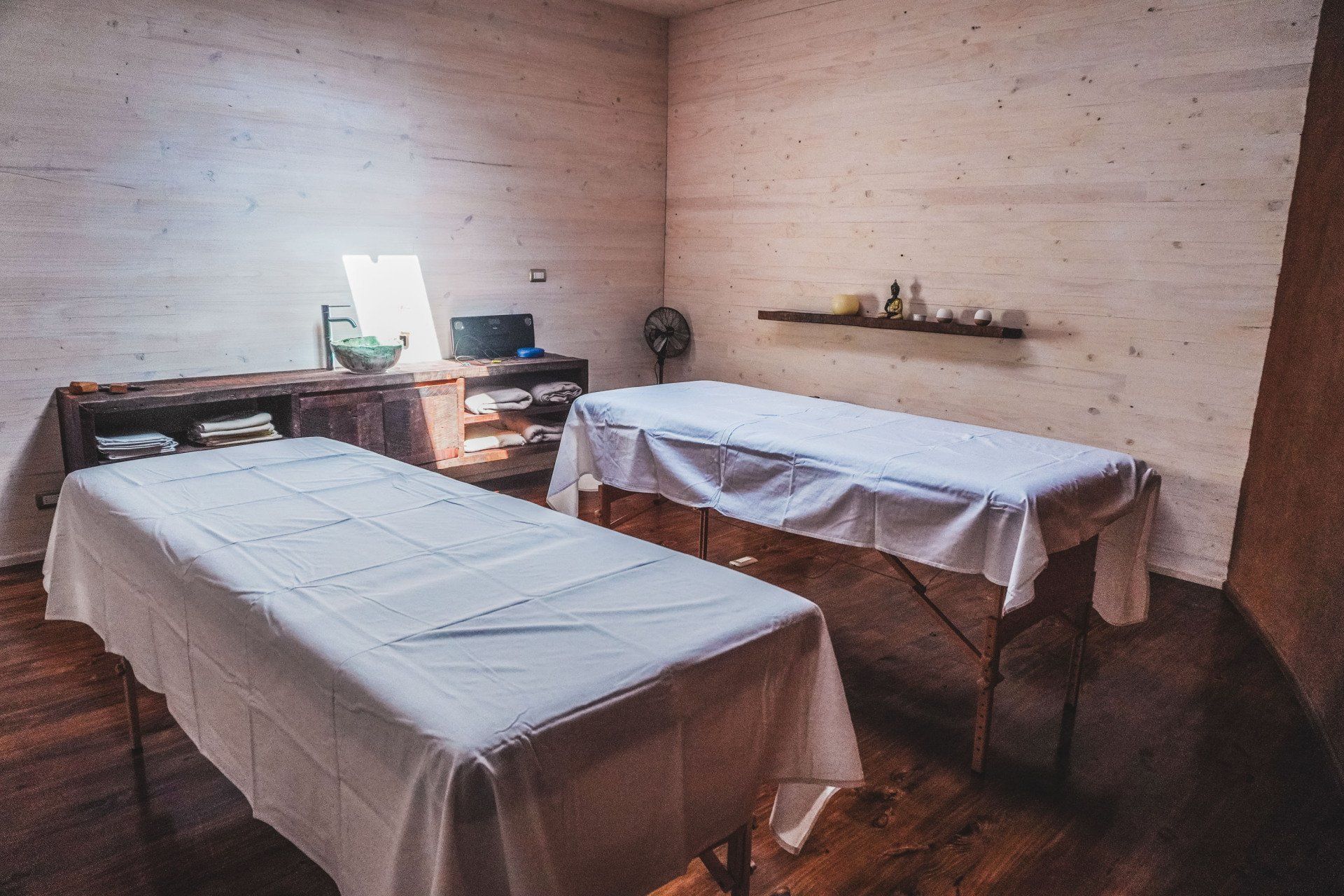 Two massage tables in a dimly lit spa room; wooden cabinets and shelves, white linens, fan, and wall decor.