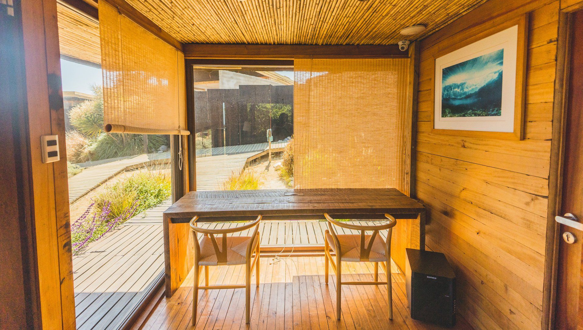 Wooden desk with two chairs, inside a cabin with outdoor view, artwork on the wall.