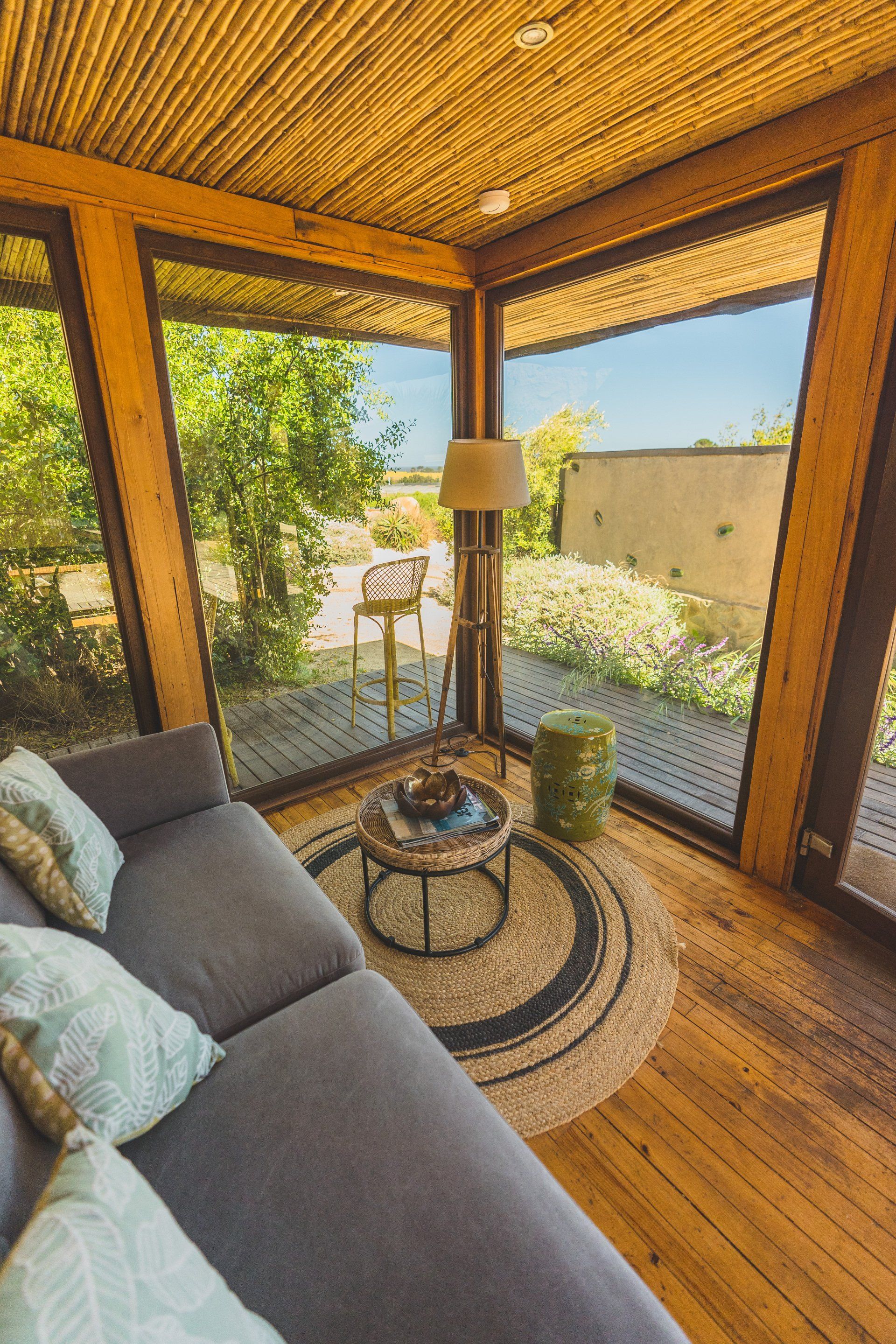 Cozy sunroom with large windows, gray sofa, round rug, and lamp; view of outdoor greenery and blue sky.