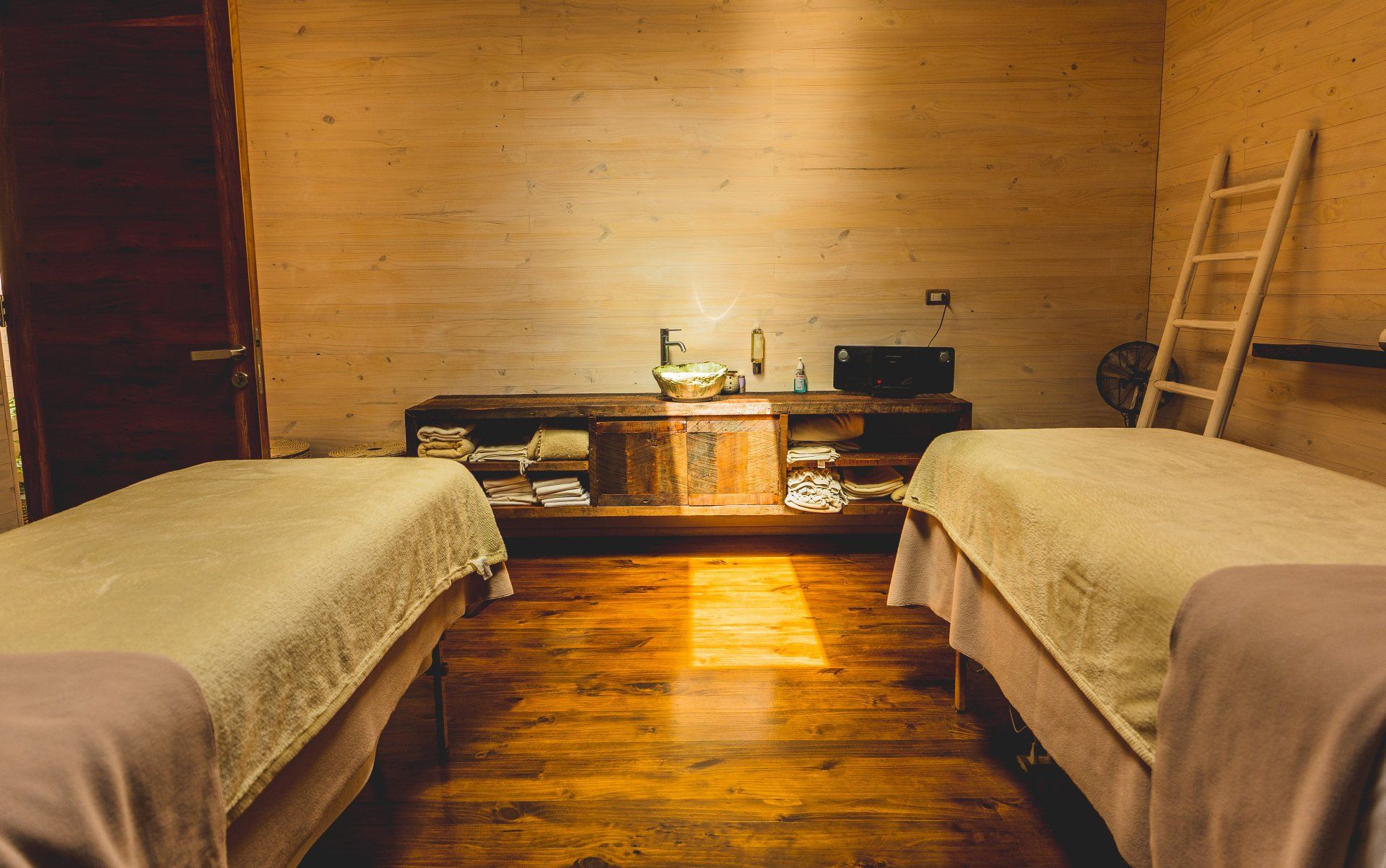 Two massage tables in a spa room with wooden floor and walls, a cabinet, and a ladder.