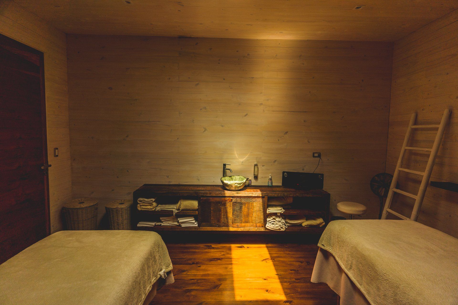 Spa room with two massage tables, wooden cabinet, and ladder against a textured wall.