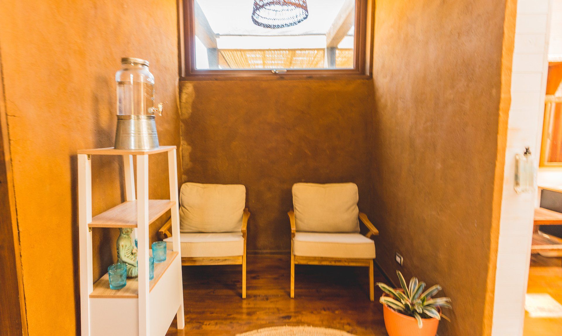 Cozy waiting area with two armchairs, earth-toned walls, wooden shelf with water dispenser, and a small plant.