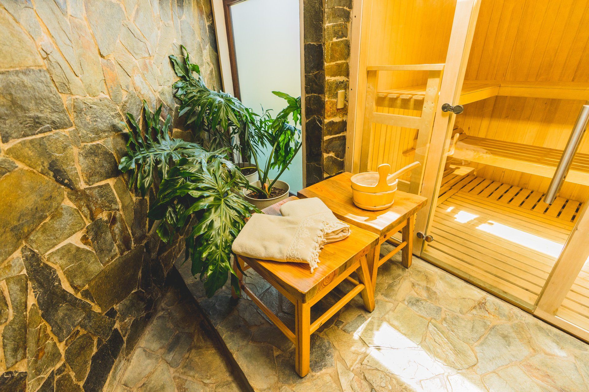 Stone-walled spa with plants, wooden stools, towels, and sauna; bright, warm light.