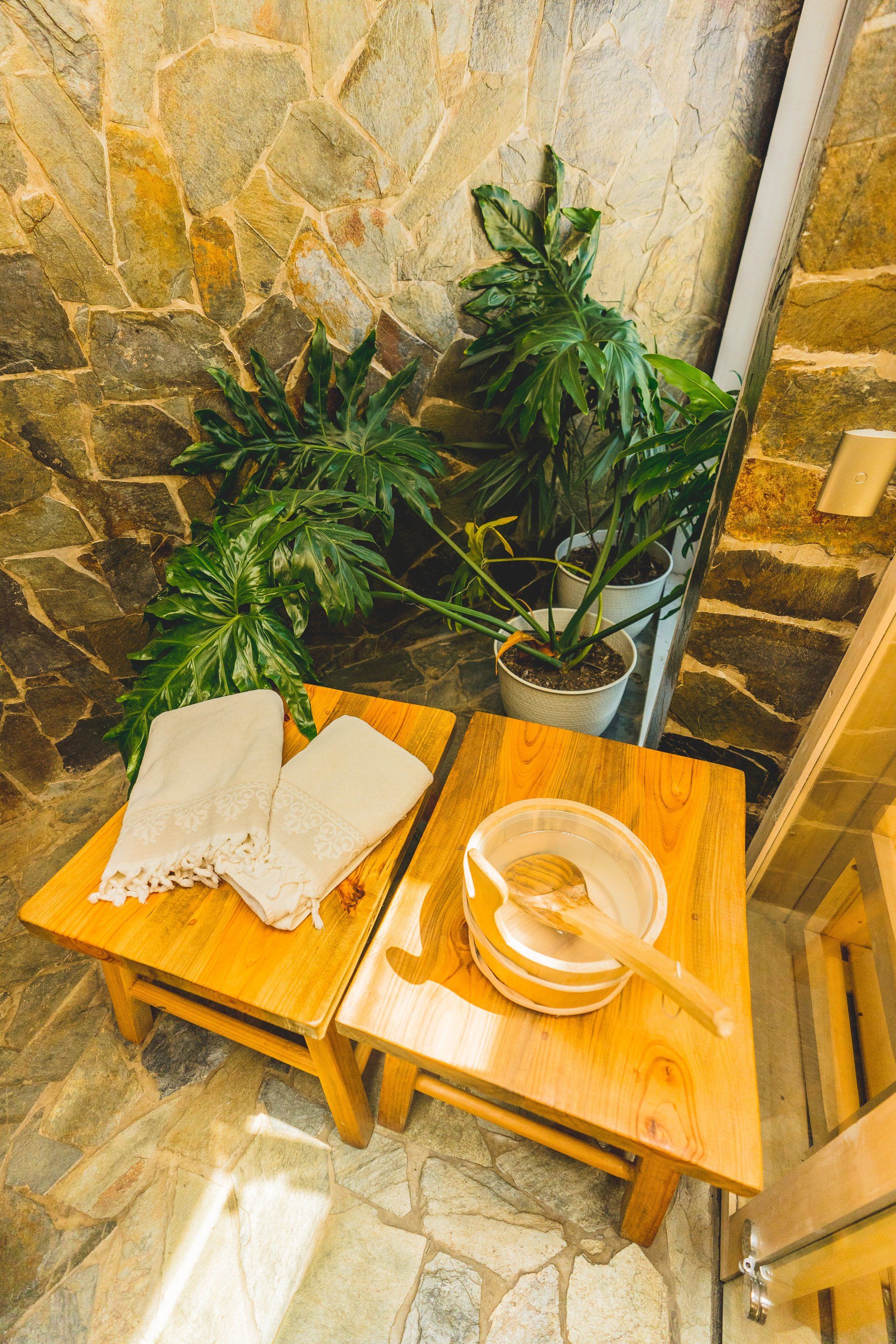 Two wooden stools with towels and a wash basin in a spa-like setting with plants. Stone wall in background.