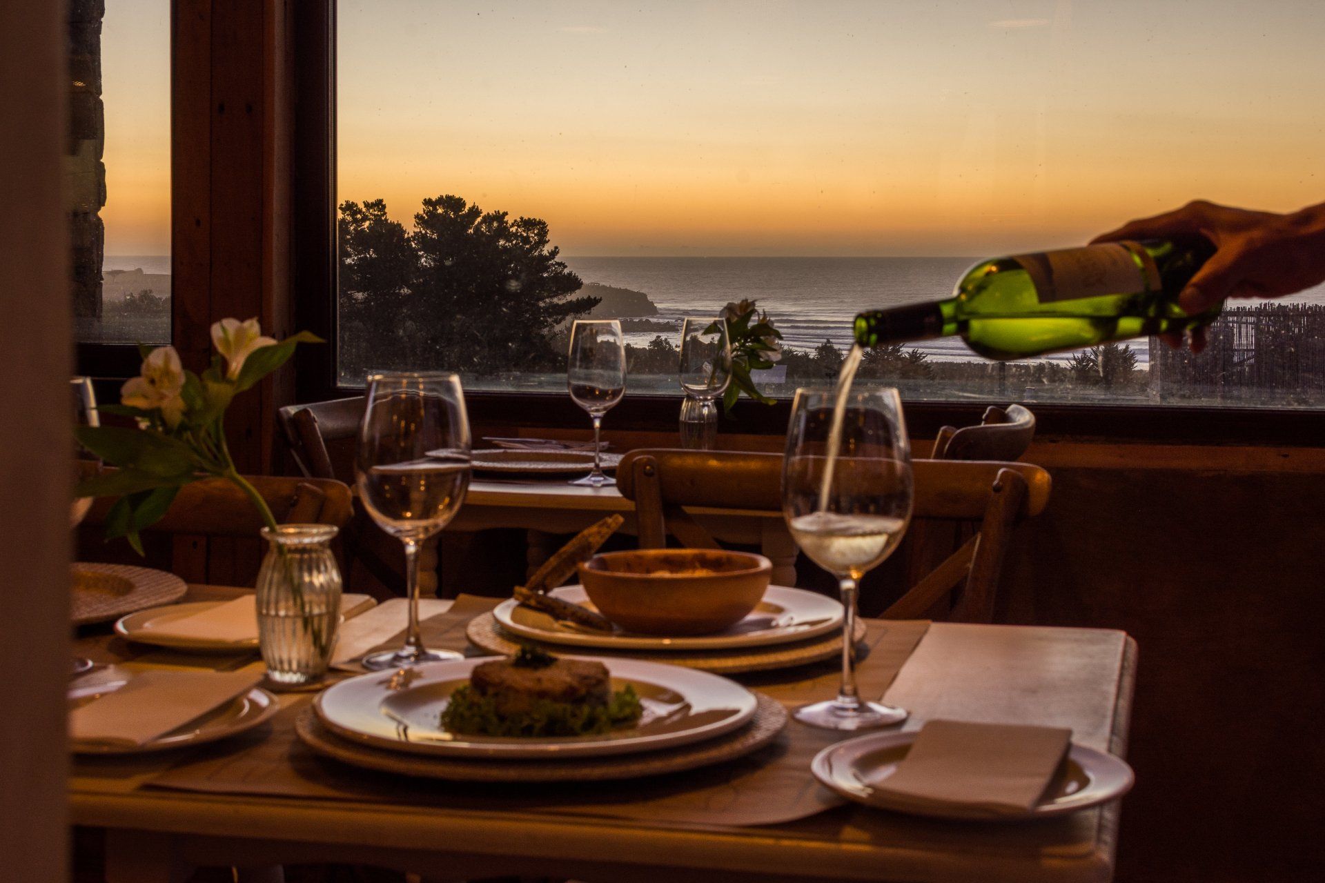 Table set for dinner with wine being poured, overlooking ocean at sunset.